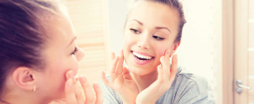 A young woman smiles while looking in the mirror. She touches her cheeks with her fingers.